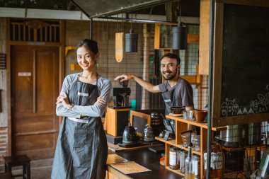 Kadın bir barista elleri çapraz dururken erkek barmen masanın içinde kahve makinesini çalıştırıyor.