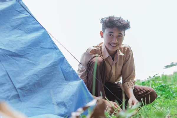 a teenage boy wearing scout clothes smiles at the camera while attaching a rope to a tent peg