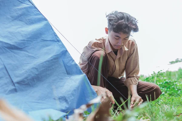 a teenage boy wearing scout clothes set up tent pegs while in the field