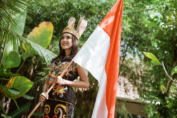 インドネシアの国旗の棒を手にしながら笑顔で立つ白昼の部族の伝統的な服を着た美しい女性 — ストック写真