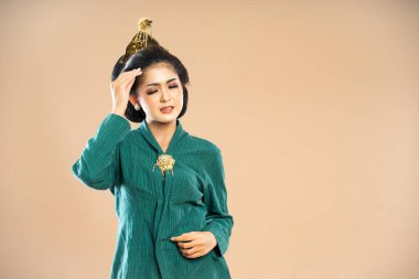 Yeşil kebaya giymiş Asyalı bir kadın gözleri kapalı duruyor ve izole bir arka planda acı içinde başını okşuyor.