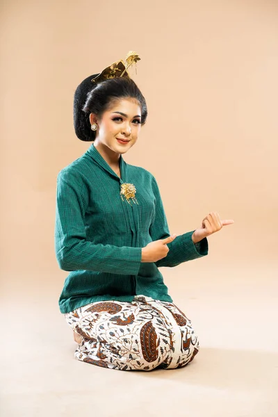 身穿绿泡菜的爪哇妇女坐在地板上 用大拇指在孤立的背景下指指着 — 图库照片