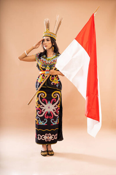 asian woman in king bibinge looking right while standing with salutation and holding the indonesian flag on isolated background