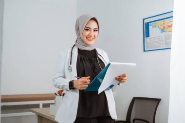 Doktor önlüğü ve gri peçeli bir kadın doktor elinde mavi bir panoyla doktor masasına yaslanıyor ve gülümsüyor.