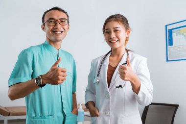 Doktor önlüklü güzel bir doktor ve hemşire üniformalı yakışıklı bir hemşire baş parmakları havada ve gülümsüyor.