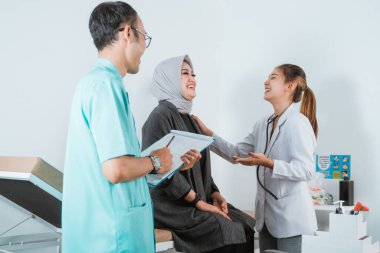 Kadın bir doktor kadın hasta ve erkek hemşire yatakta oturan kadın hasta için sağlık kontrolü yaparken birlikte gülüyorlar.