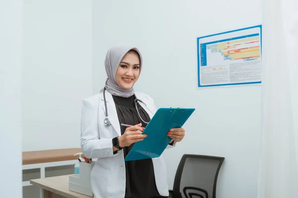 一位穿着医生外套的女医生靠着医生桌子站着 手里拿着蓝色的剪贴板 面带微笑 — 图库照片
