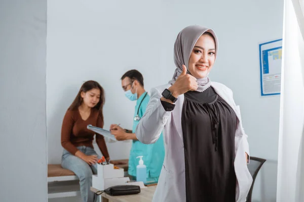 a beautiful doctor in doctors coat standing with thumb up and smiling while the nurse showing the medical check up result to the patient at the background