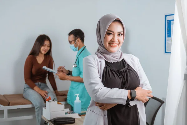a beautiful doctor in doctors coat standing with hand crossed and smiling while the nurse showing the medical check up result to the patient at the background