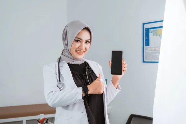 a beautiful doctor in doctors coat standing with thumb up and showing the phone at her hand