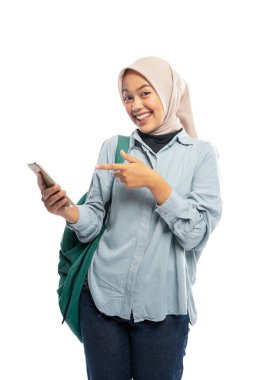 gülümseyen Asyalı kız tesettür öğrencisi beyaz arka planda cep telefonuna işaret ediyor