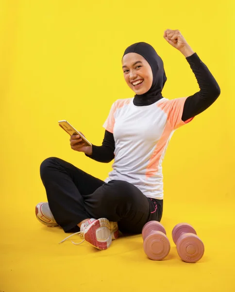 興奮アジアのイスラム教徒の女性は黄色の背景に携帯電話を保持座ってスポーツウェアを着て — ストック写真