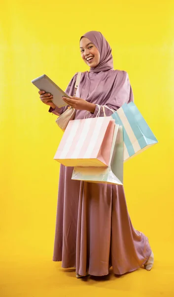 Asian woman wearing hijab dress using tablet with shopping bag on yellow background