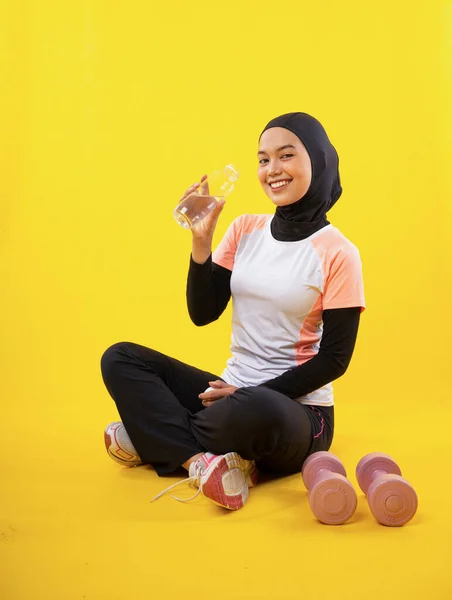 アジア系ムスリム女性を笑顔でスポーツウェアで座って水を飲みますボトルとともに黄色の背景 — ストック写真