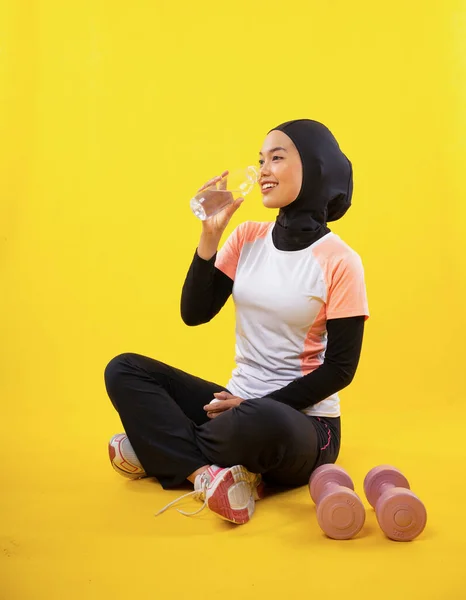 スポーツウェアのアジアのイスラム教徒の女性は黄色の背景にボトルで飲料水に座っている — ストック写真