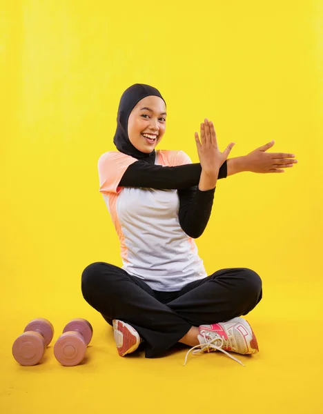 穿着运动服的快乐的亚洲穆斯林妇女坐在黄色的背景上伸展着手的肌肉 — 图库照片