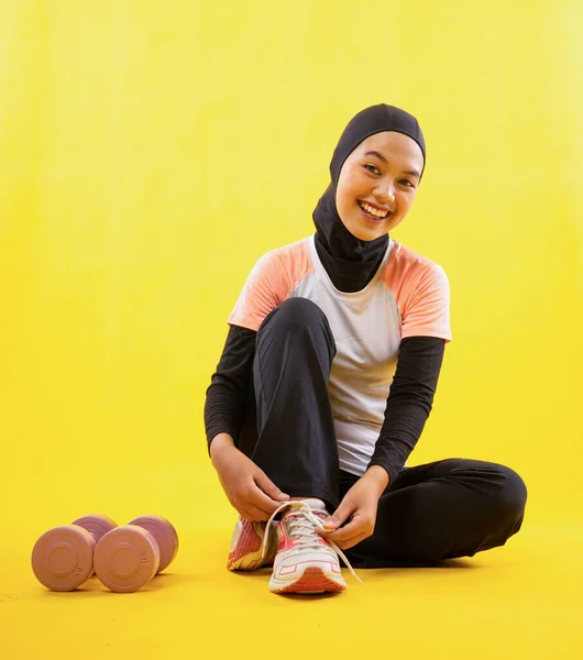 笑いイスラム教徒の女性を身に着けていますスポーツウェアは黄色の背景に靴ひもを固定座って — ストック写真