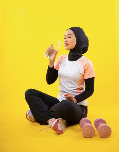 スポーツウェアのアジアのイスラム教徒の女性は黄色の背景にボトルで飲んで座っている — ストック写真