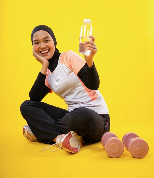スポーツウェアのアジアのイスラム教徒の女性は黄色の背景に水ボトルを示して座っています — ストック写真