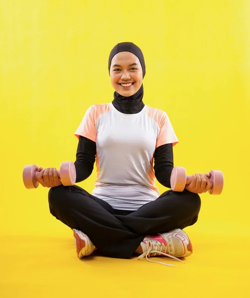 スポーツウェアを身に着けているイスラム教徒の女性は 黄色の背景にダンベルを使用してクロス脚運動に座る — ストック写真