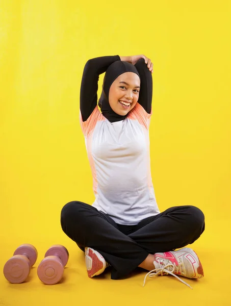 身穿运动服头戴头巾的亚洲穆斯林妇女坐在黄色背景下伸展手臂肌肉 — 图库照片
