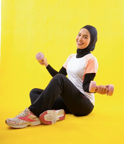 ムスリム女性でスポーツウェア座ってリラックスとともに二本の腕運ぶダンベル上の黄色の背景 — ストック写真