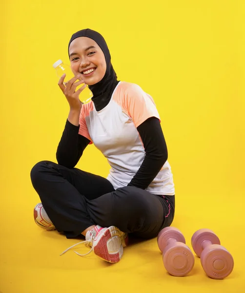 笑顔アジアのイスラム教徒女性でスポーツウェアで座っています水ボトルと黄色の背景 — ストック写真