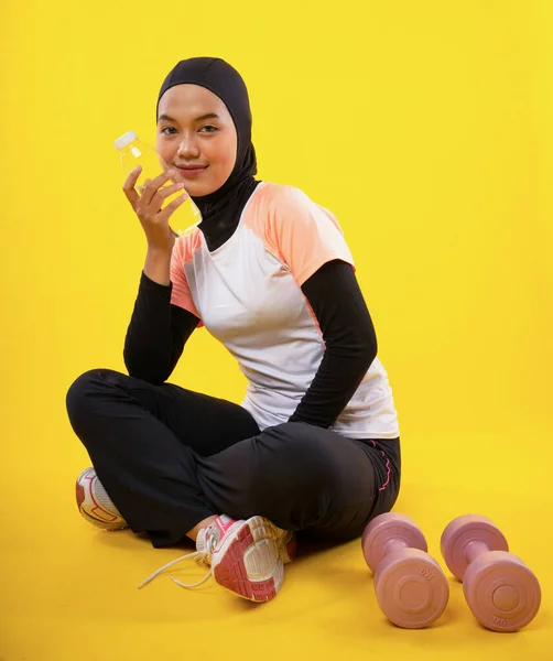 スポーツウェアのアジアのイスラム教徒の女性は黄色の背景に水ボトルを保持座っている — ストック写真
