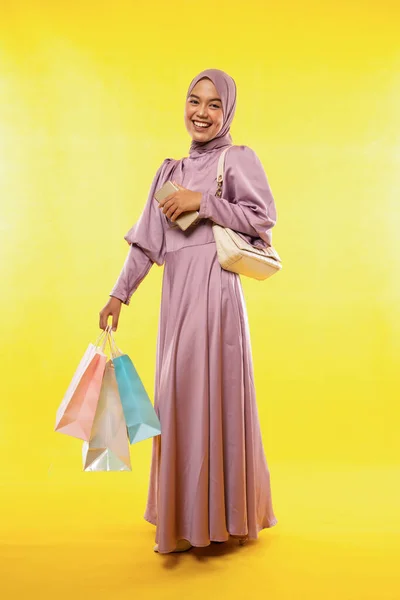Asian woman wearing color dress with hijab carrying shopping bags and mobile phone while shopping on yellow background