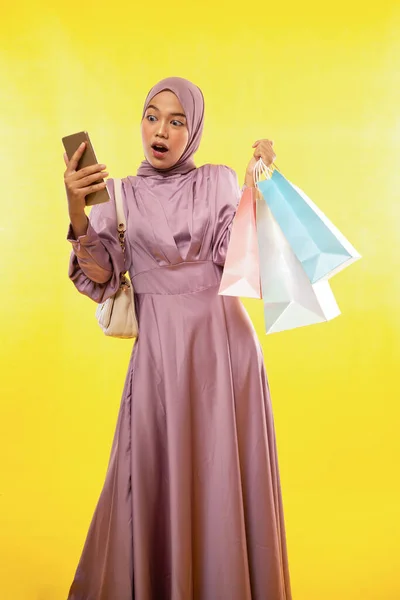 震惊的女性穆斯林站在那里看着装有黄色背景购物袋的手机 — 图库照片