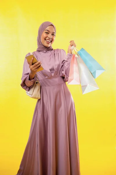 女性のイスラム教徒が携帯電話で笑顔で黄色い背景に紙袋を持っています — ストック写真