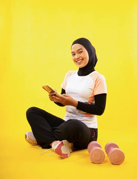 身穿运动服的亚洲穆斯林妇女双腿交叉坐着 用黄色背景的手机休息 — 图库照片