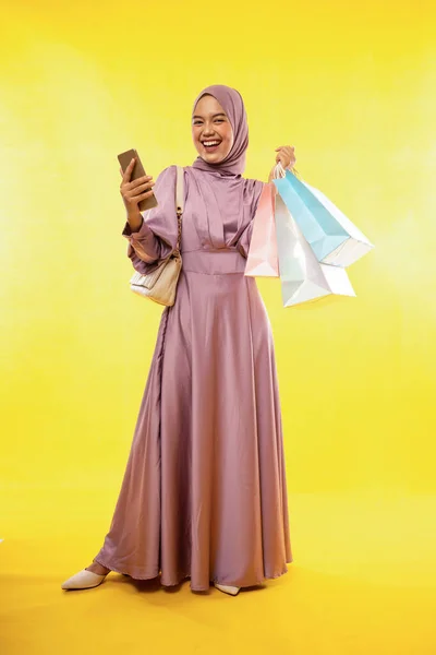 快乐的亚洲穆斯林妇女站在那里 手里拿着装有黄色背景购物袋的手机 — 图库照片