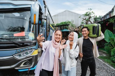 Üç genç insan gülümsüyor ve cep telefonu kameralarına poz veriyor. Otobüsün önünde selfie çekiyorlar.