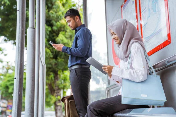 Asian man standing and veiled woman using tablet on way home from work sitting at bus stop