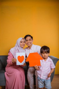 Asyalı bir aile kanepeye otururken kağıt bir aşk çizimi ve kağıt ev şekilleri gösteriyor.