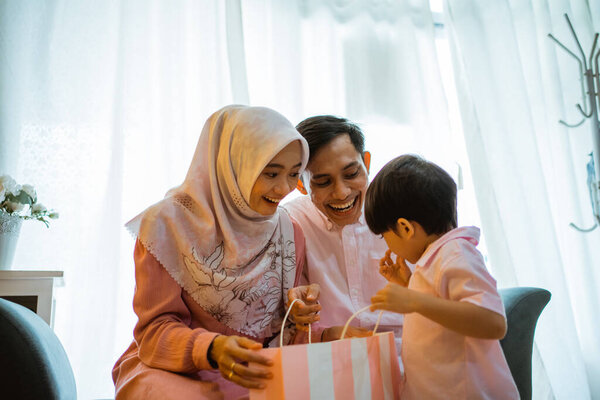 mom and son open a paper bag gift from dad during a home celebrations. Asian Muslim family concept