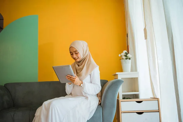 portrait of attractive Asian hijab woman sitting on sofa using digital tablet at home