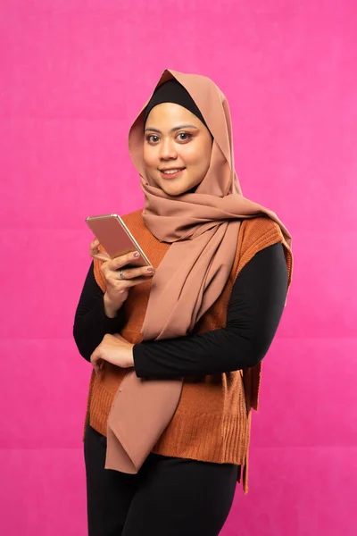 戴头巾的快乐的亚洲穆斯林女人拿着粉色背景的手机站着 — 图库照片