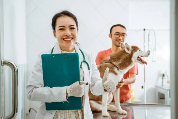 ガラスのドアの近くに顔を笑顔で立っている美しい獣医です背景にはシベリア犬とその所有者に対して親指を立てています — ストック写真