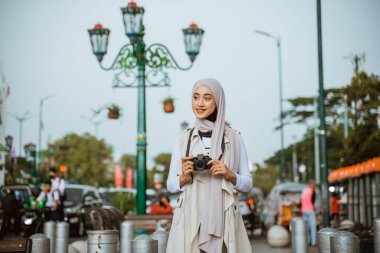 Kadın müslüman gezgin gülümseyerek kamerayı tutarken arka planda trafik sıkışıklığı olan kaldırımda duruyor.