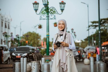 tesettürlü güzel gezgin gülümseyerek dişlerini gösteriyor ve kaldırımda dikilirken kamerayı tutuyor.