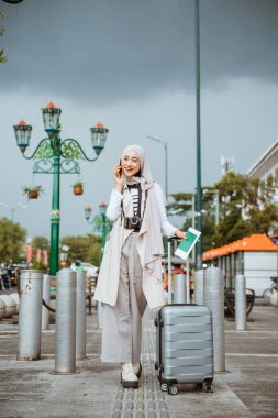 Asyalı tesettürlü gezgin, kaldırımda dururken telefonla arıyor. Bagajı ve pasaportu getir.