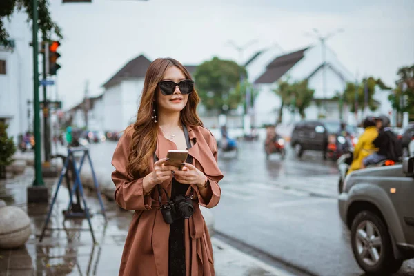 stock image female traveller with long brown hair wearing glasses and holding the phone while standing at the sidewalk