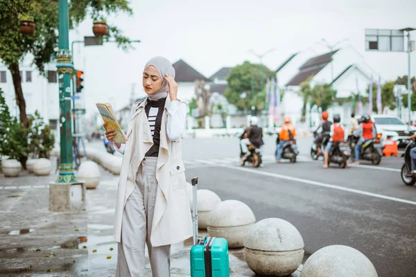 confuse asian traveller with hijab touch her head and looking at the maps while standing at the sidewalk with the traffic light at the background