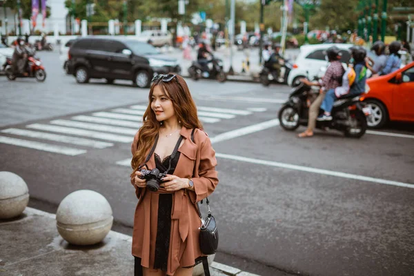 stock image female traveller with long brown hair standing at the sidewalk and holding the camera with the traffic at the background