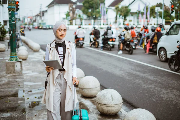 confuse asian traveller with hijab holding the digital tablet and bring the trunk while walking through the sidewalk