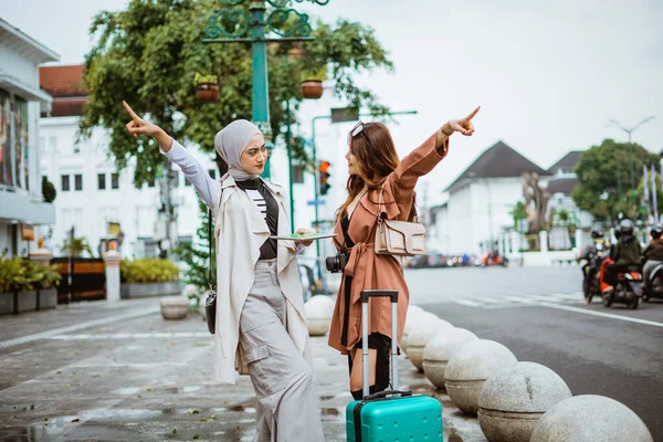two female travellers pointing to the opposite way while standing together at the sidewalk bring their travel equipment