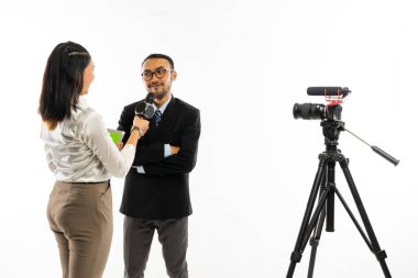 Siyah takım elbiseli yetişkin bir adam beyaz bluzlu bir kadın gazeteci tarafından sorgulanırken ellerini çapraz yapıp gülümsüyor.