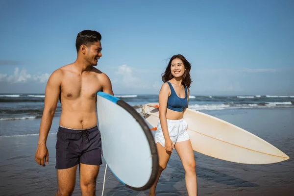 身穿泳衣的年轻夫妇在练习后背着冲浪板在蓝天背景的海滩上散步 — 图库照片
